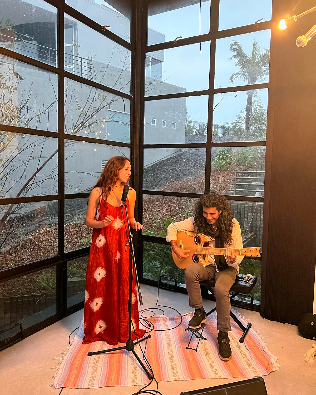 Nahti in a red dress at a microphone and Erez seated with a guitar in a glass walled room at dusk