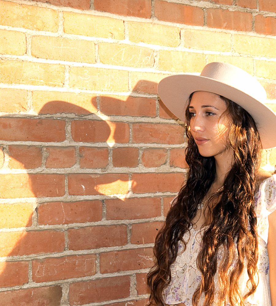 Nahti LaLuz in a pale wide brimmed hat and a floral dress against a sunlit brick wall
