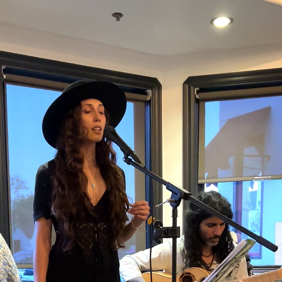 Nahti singing into a microphone in a black hat, Erez on guitar behind her