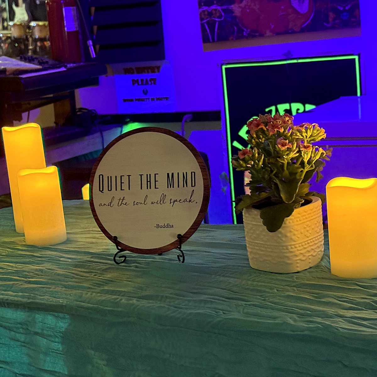 A round wooden sign reading "Quiet the mind and the soul will speak, Buddha" between candles and a flowering plant